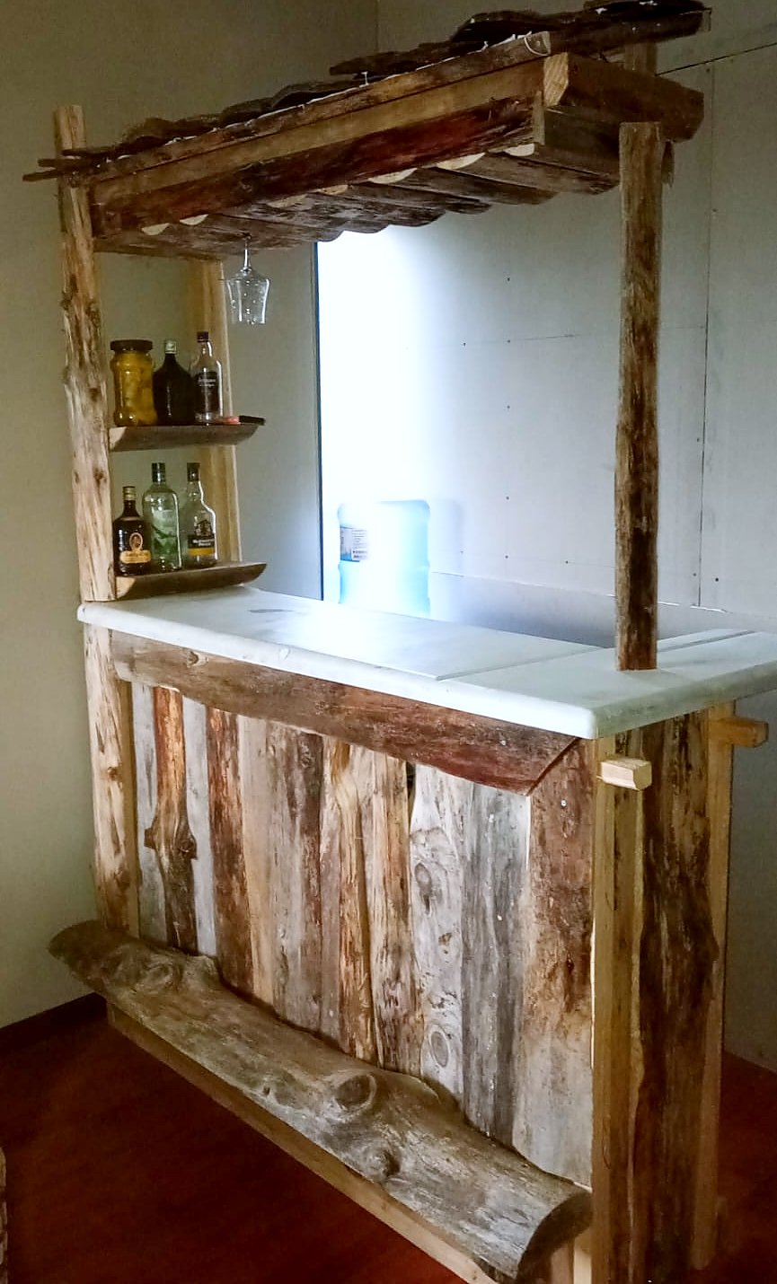 Bar de madera rústica terminado: frente de tablas de distintos tonos, mesón de cubierta blanca, repisas con botellas, copero y techo